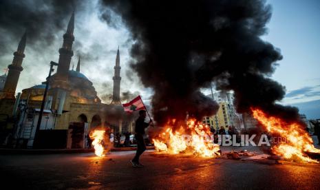  Seorang pengunjuk rasa mengibarkan bendera nasional Lebanon saat dia berjalan di depan ban yang terbakar yang memblokir jalan utama, selama protes di pusat kota Beirut, Lebanon, Rabu, 3 Maret 2021.