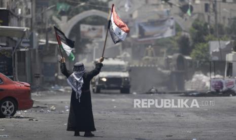 Seorang pria Palestina mengibarkan bendera Palestina dan Suriah di depan kendaraan tentara Israel selama serangan militer di kamp pengungsi Jenin, kubu militan, di Tepi Barat, 4 Juli 2023.