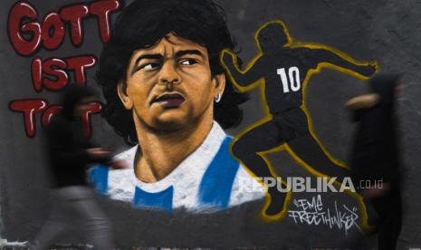 Dua orang berjalan di depan mural karya seniman Eme Freethinker untuk menghormati mendiang legenda sepak bola Argentina Diego Maradona, di dinding grafiti di Mauerpark di Berlin, Jerman, Senin, 30 November, pemain sepak bola Argentina meninggal dunia karena serangan jantung di rumahnya Rabu, 25 November, pada usia 60. 2020. Slogannya berbunyi: Tuhan adalah kematian.