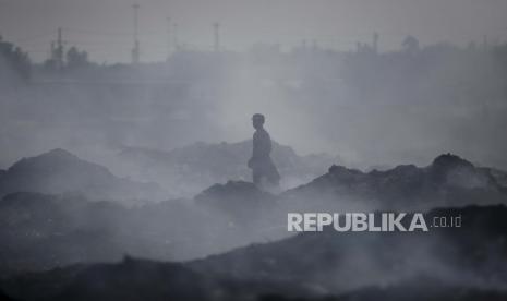 Seorang pria yang mengais bahan daur ulang untuk mencari nafkah berjalan melintasi gunung garasi di tengah asap dari pembakaran sampah.