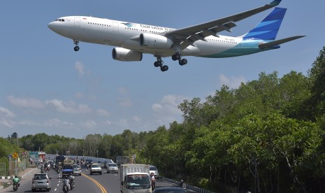 Sebuah pesawat udara terbang melintas di atas jalan raya saat bersiap mendarat di Bandara Internasional I Gusti Ngurah Rai, Denpasar, Bali. Karantina Pertanian Denpasar menyiapkan karpet disinfektan untuk mencegah masuk, keluar dan tersebarnya Penyakit Mulut dan Kuku (PMK) di Bandara Internasional I Gusti Ngurah Rai Bali.