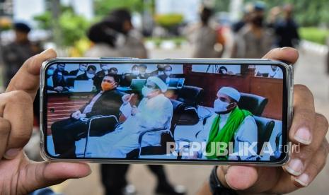 Lewat layar telepon genggam, pendukung menonton sidang Habib Rizieq Shihab di luar Pengadilan Negeri Jakarta Timur, Kamis (27/5). 