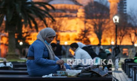 Orang-orang berbuka puasa dengan latar belakang Masjid Sultan Ahmed yang ikonik, lebih dikenal sebagai Masjid Biru, dihiasi dengan lampu dan slogan bertuliskan Ramadhan adalah cinta, menandai bulan Ramadhan, di distrik bersejarah Sultan Ahmed di Istanbul, Turki,  Selasa (13/4).