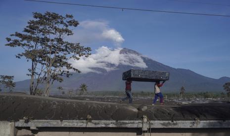 Para pria membawa kasur untuk dibawa ke tempat penampungan sementara saat mengumpulkan barang-barang dari rumahnya yang terkena dampak erupsi Gunung Semeru di Desa Sumberwuluh, Lumajang, Jawa Timur, Indonesia, Selasa, 6 Desember 2022. Gunung Semeru, latar belakang, memuntahkan kolom abu tebal lebih dari 1.500 meter (hampir 5.000 kaki) ke langit pada hari Minggu. Desa-desa dan kota-kota terdekat diselimuti abu yang jatuh, menghalangi sinar matahari, tetapi tidak ada korban yang dilaporkan. 