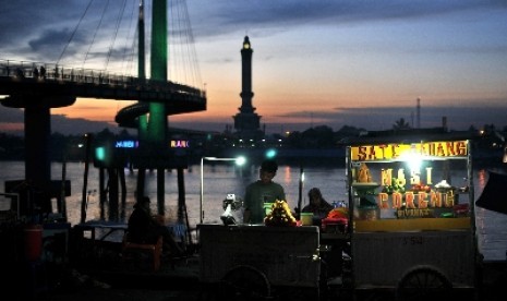 Pedagang makanan dan minuman menunggu calon pembeli di kawasan Tanggo Rajo dengan latar belakang jembatan pedestrian Gentala Arasy dan Sungai Batanghari di Jambi.