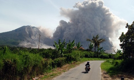 Pengendara sepeda motor melintas dengan latar belakang Gunung Sinabung yang tengah meluncurkan awan panas guguran kubah lava Gunung Sinabung di Karo, Sumatera Utara, Rabu (24/6).