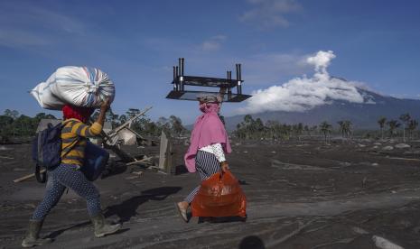 Perempuan membawa barang bawaan yang mereka kumpulkan dari rumah mereka yang terkena dampak letusan Gunung Semeru di Desa Sumberwuluh, Lumajang, Jawa Timur, Indonesia, Selasa, 6 Desember 2022. Gunung Semeru, latar belakang, memuntahkan abu tebal lebih dari 1.500 meter (hampir 5.000 kaki) ke langit hari Minggu. Desa-desa dan kota-kota terdekat diselimuti abu yang jatuh, menghalangi sinar matahari, tetapi tidak ada korban yang dilaporkan. 