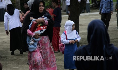 Seorang ibu berjalan sambil menggendong balitanya mendampingi anaknya pergi ke sekolah.