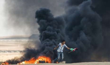 Seorang pengunjuk rasa Palestina berlari melewati ban yang terbakar sambil memegang bendera Palestina. PBB mempertahankan konsensus internasional tentang konflik Israel-Palestina. Ilustrasi.