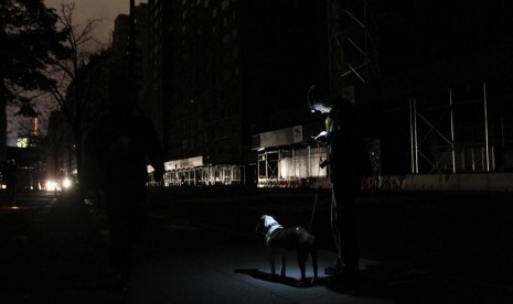   Seorang warga dengan anjing peliharaannya berdiri di tengah jalan yang gelap gulita akibat terputusnya aliran listrik di New York, Rabu (31/10).      (Tina Fineberg/AP)