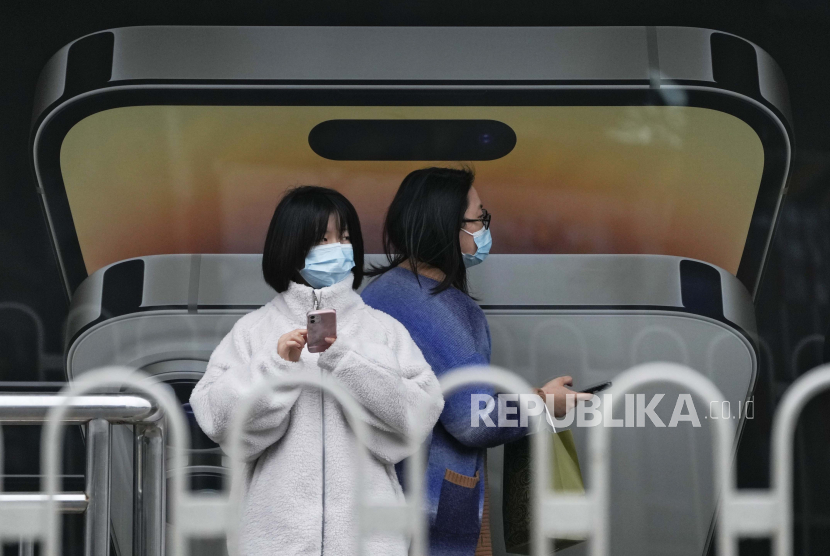 Seorang wanita yang mengenakan masker berjalan dengan yang lain menggunakan iPhone saat mereka menunggu bus di halte.
