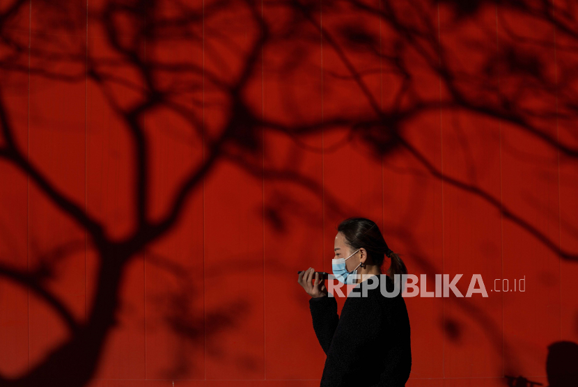 Seorang wanita yang mengenakan masker untuk membantu mengekang penyebaran pembicaraan virus corona di smartphone-nya saat dia berjalan melewati bayangan pohon di atas truk yang diparkir di jalan di Beijing, Senin, 7 Desember 2020.