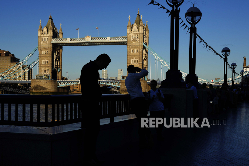 Seorang turis memeriksa ponselnya ketika yang lain mengambil foto Tower Bridge di tepi selatan sungai Thames, di London, Inggris.