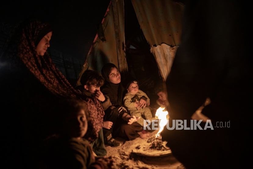 Keluarga Okasha, pengungsi dari Jalur Gaza utara, menghangatkan diri di dekat api unggun di luar tenda, dekat perbatasan yang memisahkan Jalur Gaza dan Mesir, di Rafah, Jalur Gaza selatan (27/2/2024).