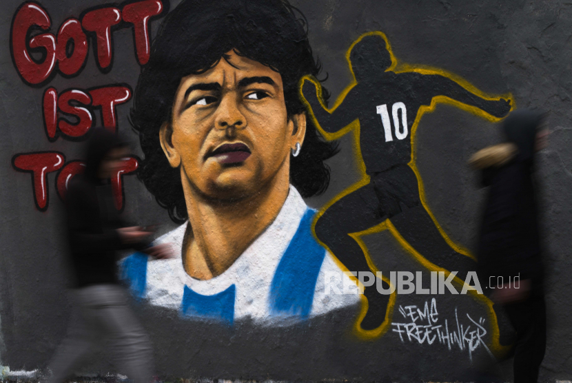 Dua orang berjalan di depan mural karya seniman Eme Freethinker untuk menghormati mendiang legenda sepak bola Argentina Diego Maradona, di dinding grafiti di Mauerpark di Berlin, Jerman, Senin, 30 November, pemain sepak bola Argentina meninggal dunia karena serangan jantung di rumahnya Rabu, 25 November, pada usia 60. 2020. Slogannya berbunyi: 