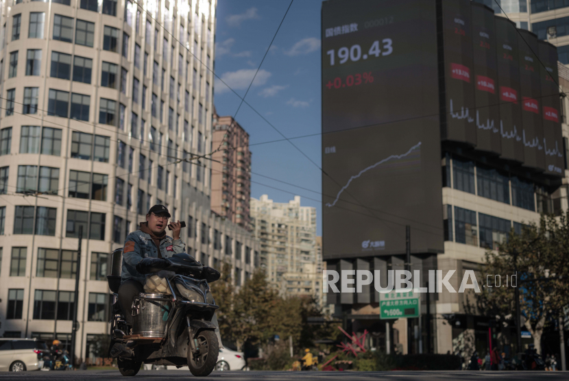  Pria mengendarai sepeda motor di jalan di sebelah layar besar yang menampilkan data bursa saham terbaru di Shanghai, Cina, 19 November 2021. Saham-saham Hong Kong ditutup pada level terendah satu bulan dan saham China jatuh pada perdagangan Senin (6/2/2023). 