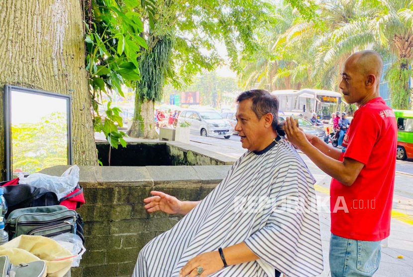 Tukang cukur rambut di jalur pedestrian Sistem Satu Arah (SSA) Kota Bogor, Idrus (49 tahun) sedang melayani pelanggannya, Sabtu (18/3/2023). 
