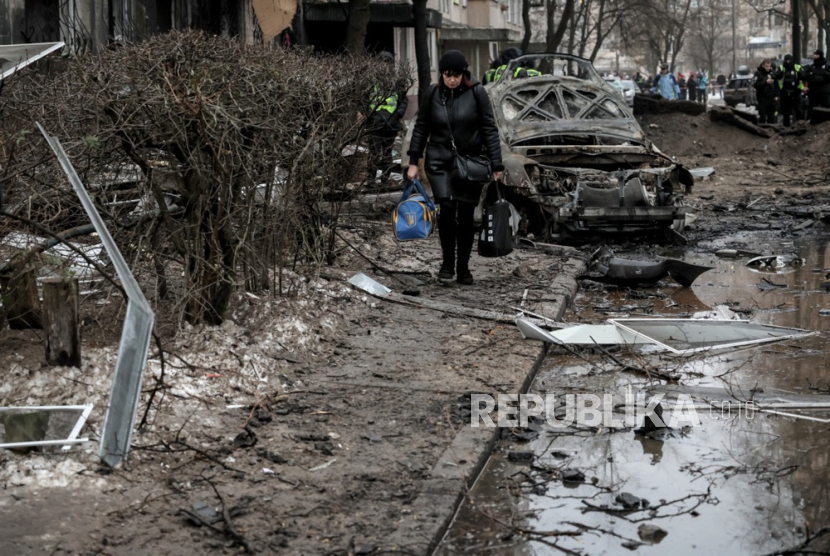 Seorang warga setempat yang membawa barang-barangnya berjalan di antara puing-puing melewati mobil yang hancur di lokasi bangunan tempat tinggal yang rusak akibat serangan rudal di Kyiv, Ukraina. 