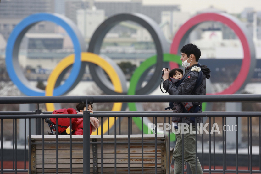  Orang-orang yang memakai masker wajah untuk melindungi dari penyebaran virus corona berdiri di tepi pantai Odaiba saat cincin Olimpiade terlihat di latar belakang di Tokyo, Selasa, Januari. 26, 2021.