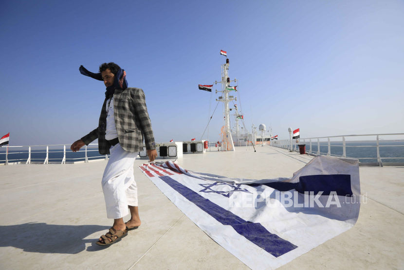 Seorang warga Yaman melewati spanduk bergambar bendera Israel dan AS di dek kapal kargo Galaxy Leader, yang disita oleh Houthi di lepas pantai pelabuhan Al-Salif di Laut Merah di provinsi Hodeidah, Yaman, Selasa (5/12/2023).