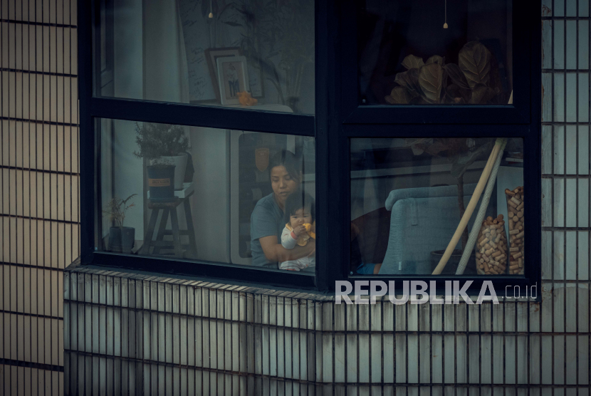 Seorang wanita dengan bayi yang dikarantina melihat melalui jendela apartemen mereka, di tengah penguncian penuh Covid-19 kota di Shanghai, Cina, 27 April 2022. 