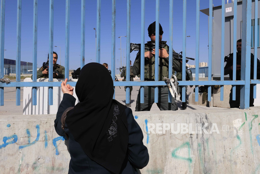 Seorang wanita Palestina bereaksi di depan petugas bersenjata Israel yang berdiri di belakang pagar di pos pemeriksaan Israel di Qalandia, antara Tepi Barat dan Yerusalem, (5/4/2024). 