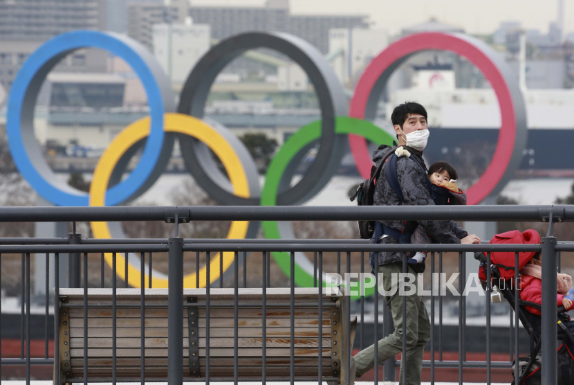  Orang-orang yang memakai masker wajah untuk melindungi dari penyebaran virus corona berjalan di tepi pantai Odaiba saat lingkaran Olimpiade terlihat di latar belakang di Tokyo, Selasa, Januari. 26, 2021.