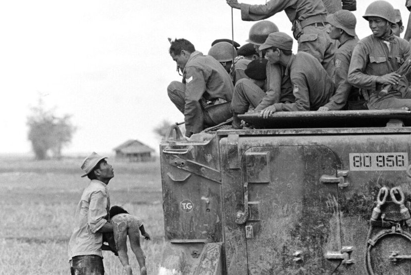 Seorang ayah menggendong jenazah anaknya saat Penjaga Tentara Vietnam Selatan melihat ke bawah dari kendaraan lapis baja mereka, 19 Maret 1964. Anak itu terbunuh saat pasukan pemerintah mengejar gerilyawan ke sebuah desa dekat perbatasan Kamboja.