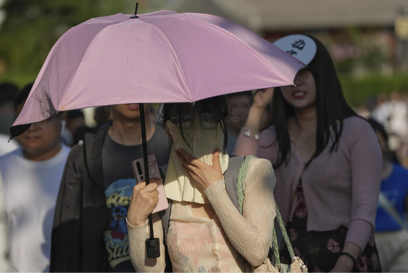 Seorang warga Cina menggunakan masker muka dan payung di tengah cuaca panas, di Beijing, Ahad (16/6/2024). Cina dilanda dua cuaca ekstrem secara bersamaan. 