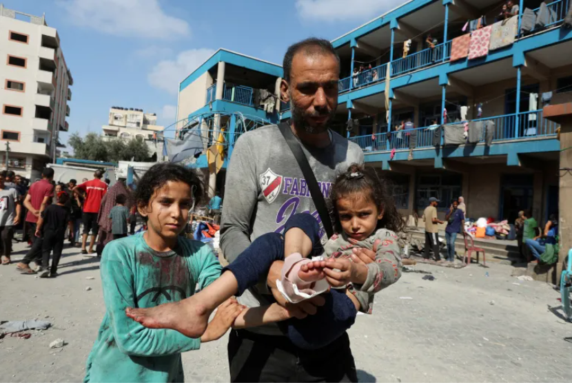 Seorang pria Palestina membawa seorang anak yang terluka setelah serangan terhadap sekolah PBB yang menampung para pengungsi, Ahad (14/7/2024).