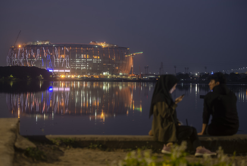 Dua warga duduk di tepi Waduk Cincin dengan latar belakang pembangunan Jakarta International Stadium (JIS) di Papanggo, Tanjung Priok, Jakarta Utara, Rabu (9/3/2022). Turnamen International Youth Championship (IYC) 2021 akan digelar di Jakarta International Stadium (JIS) pada 13-19 April 2022.