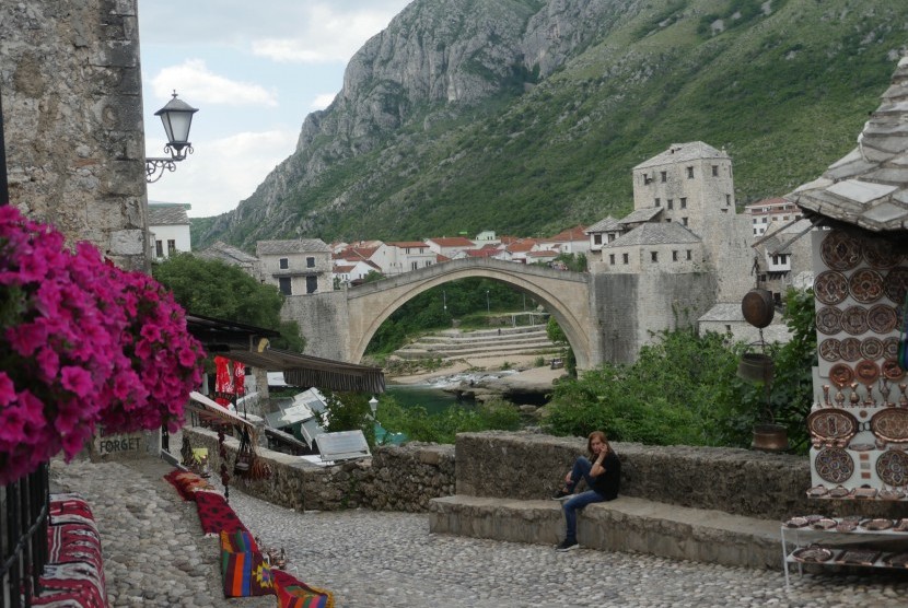 Jembatan Mostar beserta pemandangan pegunungan dan bunga berwarna merah.