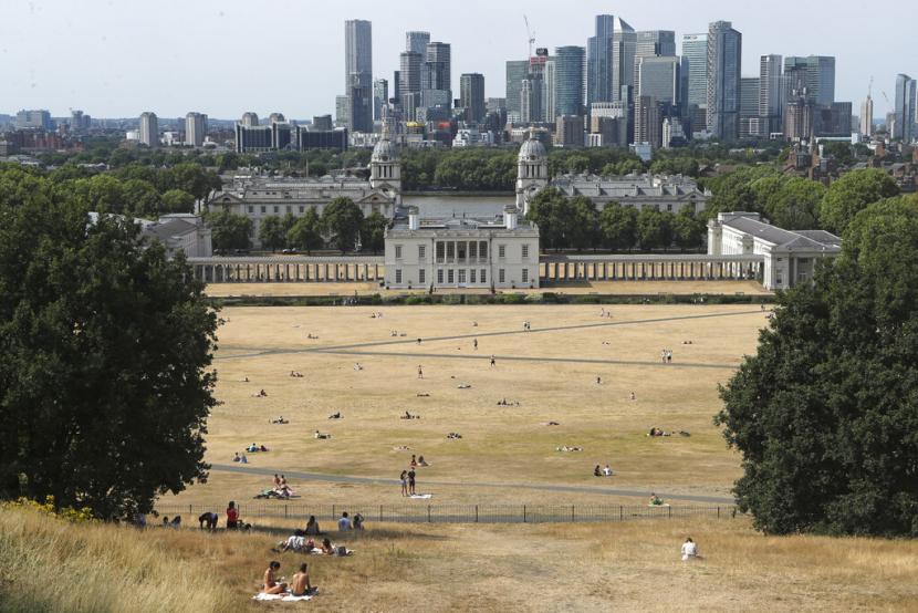 Orang-orang duduk di rerumputan yang terpanggang matahari di Greenwich Park dengan latar belakang museum Maritim dan distrik keuangan Canary Wharf di London, Minggu 17 Juli 2022. Badan cuaca Inggris telah mengeluarkan 