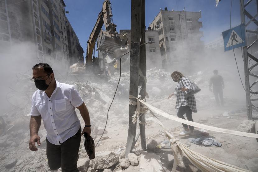 Pejalan kaki melewati awan debu saat peralatan konstruksi berat digunakan untuk menyaring puing-puing untuk menemukan barang-barang berharga sebelum diangkut dari lokasi bangunan yang hancur dalam serangan udara sebelum gencatan senjata yang menghentikan perang 11 hari antara Hamas di Gaza. 