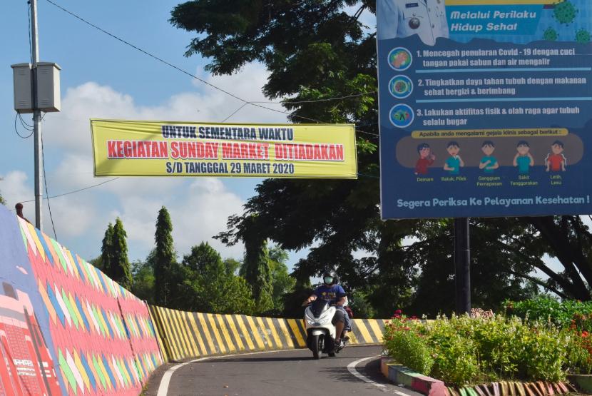Pengendara melintas di bawah spanduk bertuliskan pengumuman peniadaan kegiatan Pasar Minggu di Taman Lalu Lintas Bantaran Sungai Madiun, Ahad (22/3/2020).
