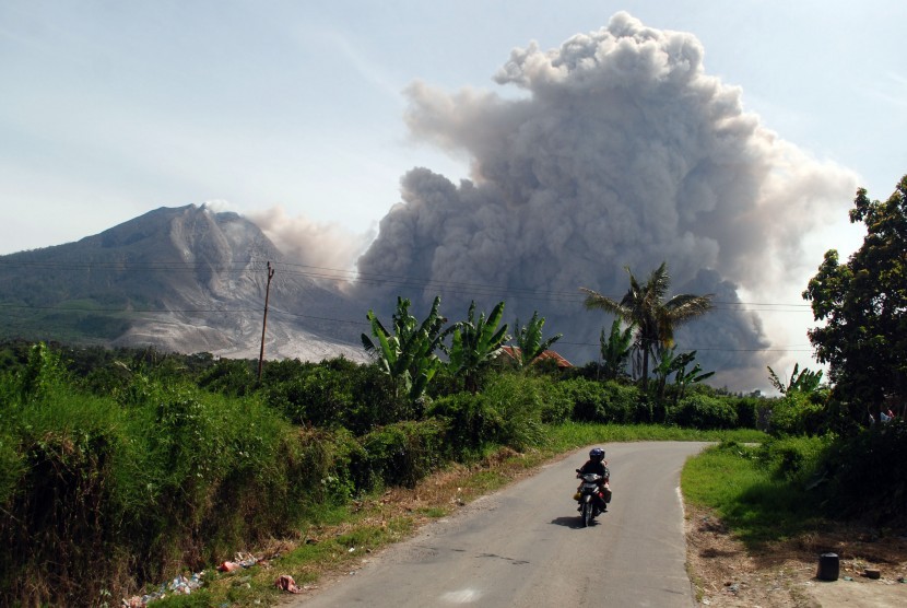 Pengendara sepeda motor melintas dengan latar belakang Gunung Sinabung yang tengah meluncurkan awan panas guguran kubah lava Gunung Sinabung di Karo, Sumatera Utara, Rabu (24/6).