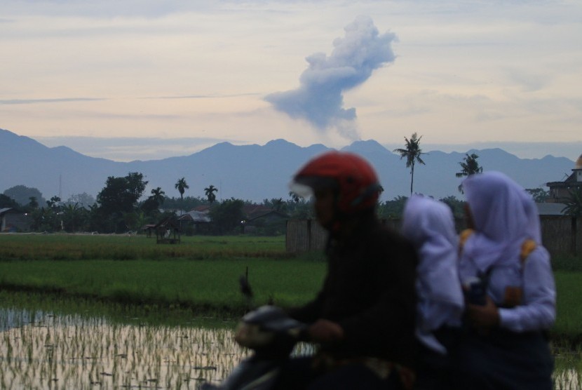 Pengendara sepeda motor melintas dengan latar belakang Gunung Sinabung menyemburkan material vulkanik tampak dari kawasan Sunggal (70 km dari Sinabung), Deli Serdang, Sumatera Utara, Selasa, (19/7). 