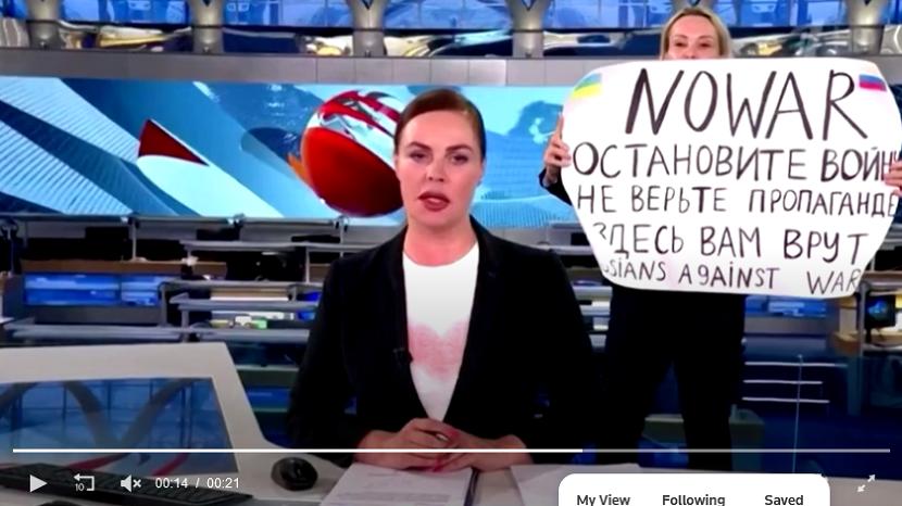 Pengunjuk rasa mengangkat sebuah kertas bertuliskan anti-perang di belakang presenter yang sedang melakukan siaran langsung di saluran televisi pemerintah Rusia, Channel One