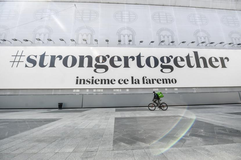  Petugas pengantar makanan melewati spanduk bertuliskan Stronger Together, kita semua akan melewatnya di Milan, Italia, Ahad (5/4).