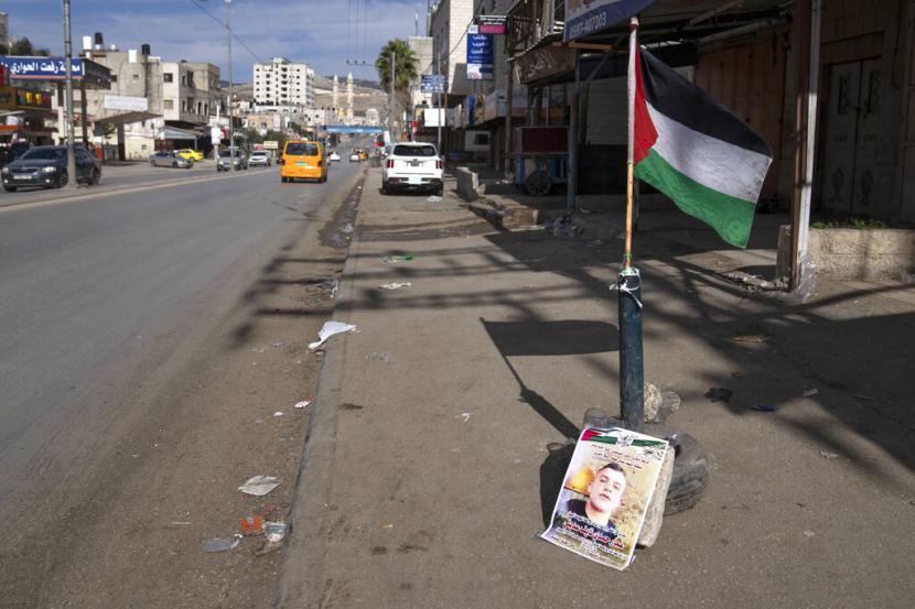 Sebuah bendera Palestina dan poster yang menunjukkan Ammar Adili, 22, yang ditembak dan dibunuh oleh seorang petugas polisi perbatasan Israel pada hari Jumat, menandai lokasi insiden tersebut, di jalan raya utama di mana toko-toko tutup dalam pemogokan umum, di Tepi Barat. kota Hawara, selatan Nablus, Sabtu, 3 Desember 2022.