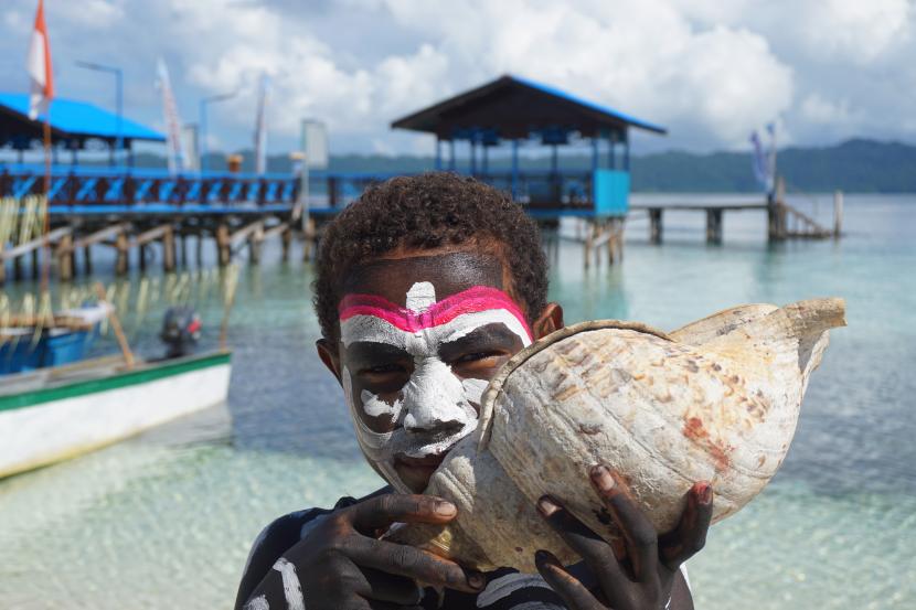 Seorang anak kampung Arborek meniup kulit Bia alat musik khas Papua di pantai Kampung Arborek, Kabupaten Raja Ampat, Papua Barat. Pemerintah kabupaten Raja Ampat, Provinsi Papua Barat, berharap bantuan infrastruktur pada destinasi wisata dari kementerian utamakan air bersih.