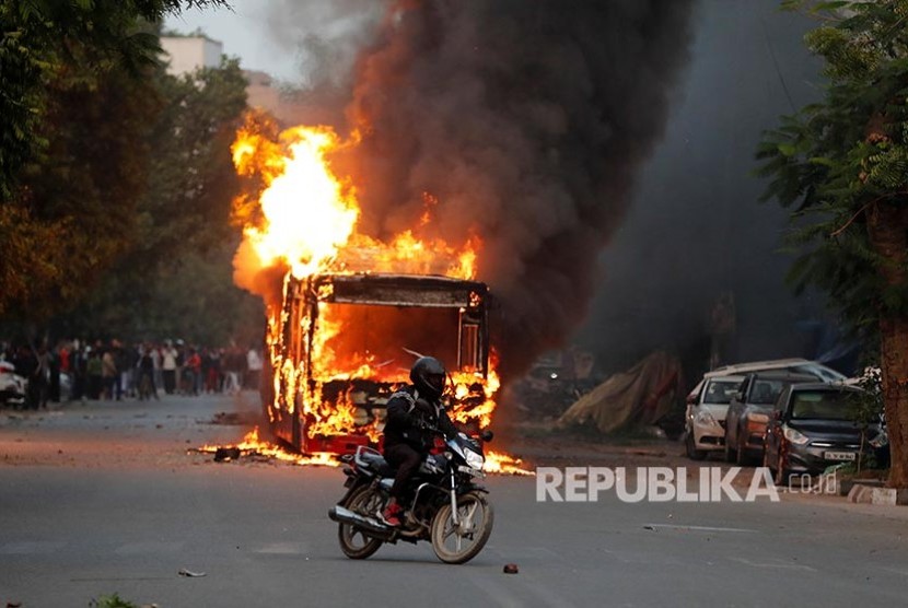 Seorang pengendara melintasi bus yang terbakar saat maasa berunjukrasa penentang Revisi UU Kewarganegaraan India di New Delhi, India, Ahad (15/12)
