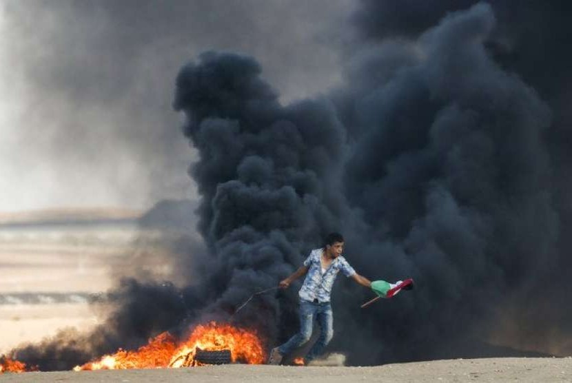 Seorang pengunjuk rasa Palestina berlari melewati ban yang terbakar sambil memegang bendera Palestina.