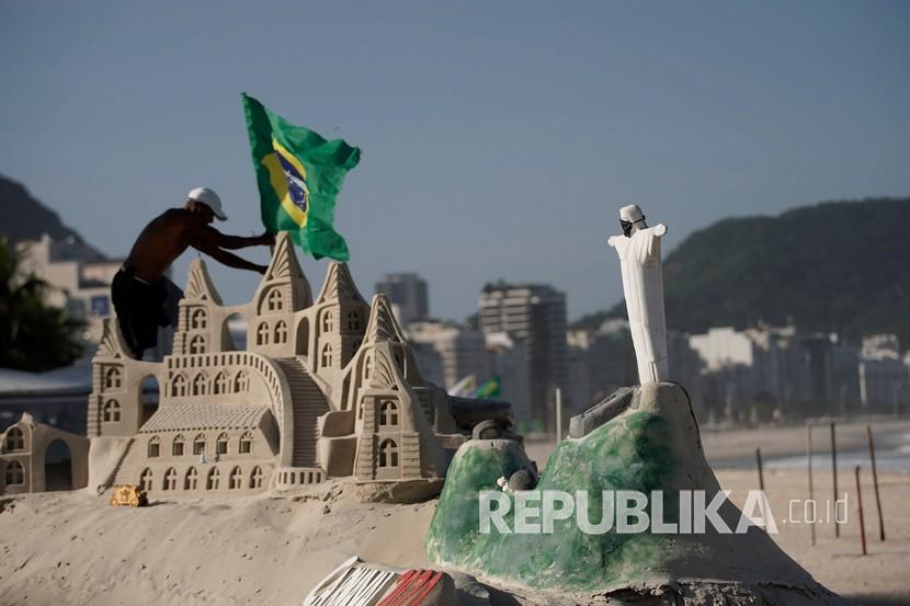 Seorang pria meletakan bendera Brasil di sebelah patung pasir Christ the Redeemer yang merupakan ikon di Rio de Janerio yang memakai masker.