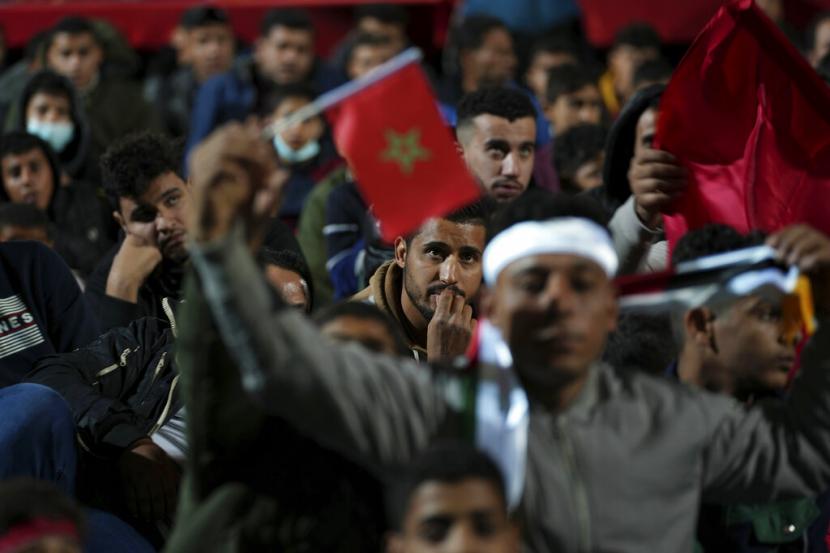Seorang pria Palestina mengibarkan bendera Maroko ketika yang lain menonton siaran langsung pertandingan sepak bola semifinal Piala Dunia antara Maroko dan Prancis yang dimainkan di Qatar, di stadion kotamadya di kamp pengungsi Rafah, Jalur Gaza Selatan, Kamis (15/12) dini hari WIB.
