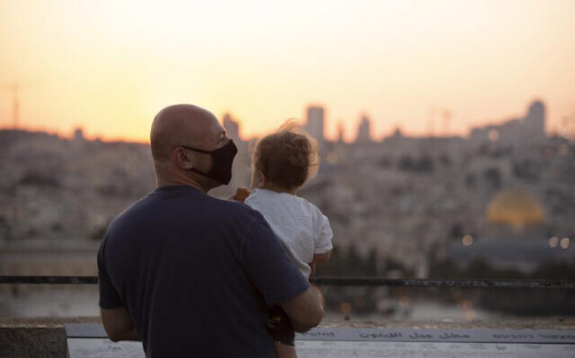 Seorang pria yang mengenakan maske untuk mengekang penyebaran virus korona menggendong seorang anak di depan Bukit Zaitun saat mereka menyaksikan matahari terbenam di balik Kubah Batu di Yerusalem, pada 2 September 2020. r