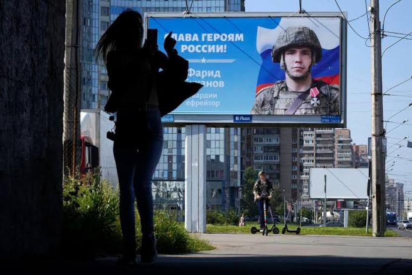 Seorang wanita menggunakan ponselnya saat dia berjalan melewati papan iklan dengan potret seorang tentara Rusia dan kata-kata 