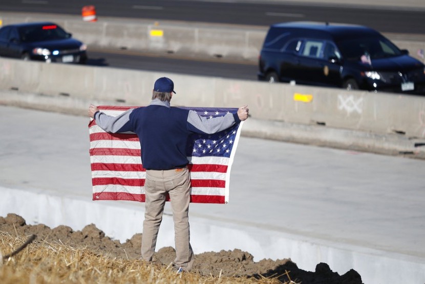 Seorang warga Colorado, Amerika Serikat (AS) membentangkan bendera AS saat kendaraan yang membawa jenazah anggota polisi korban penembakan melintas, Ahad (31/12).