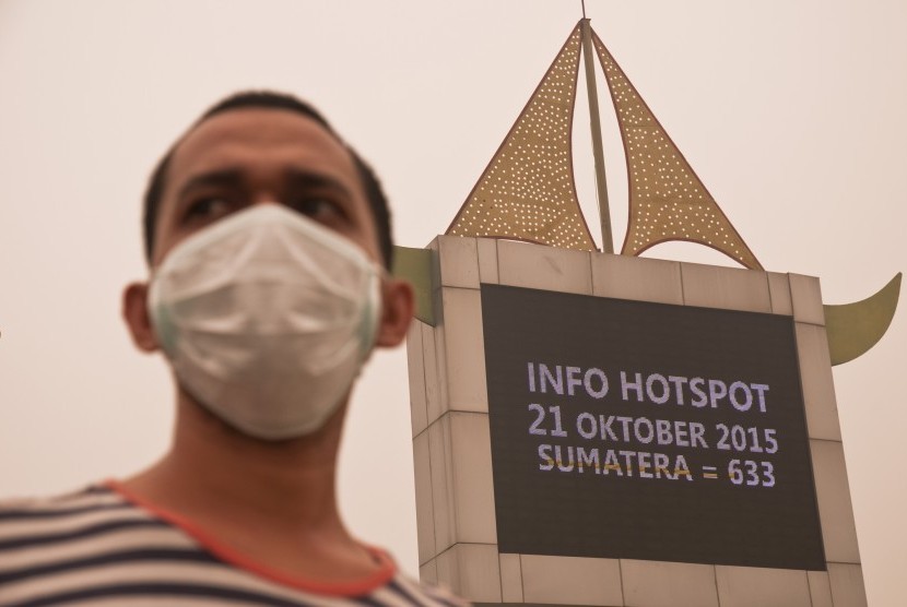 Seorang warga mengenakan masker dengan latar belakang papan informasi jumlah titik panas atau 