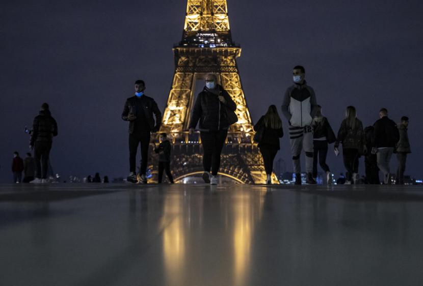 Warga Paris mengenakan masker berjalan di belakang Menara Eiffel. Kenaikan kasus Covid-19 sebabkan pemerintah Prancis kembali memberlakukan jam malam tekan laju kasus.    